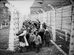 Wearing adult-size prisoner jackets, child survivors of Auschwitz are led by relief workers and Soviet soldiers through a narrow passage between two barbed-wire fences.