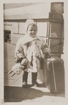 A young Jewish DP child wearing a fur coat and clutching a doll, poses next to a suitcase as her family prepares to depart from the Lampertheim displaced persons camp for the United States.