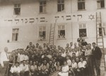Members of the Kibbutz Buchenwald hachshara are gathered outside their barracks, beneath a banner bearing their name in Hebrew.