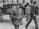 A Jew wearing a yellow star walks along a street in Theresienstadt just ahead of a man pulling a wagon of bread.