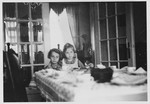 Two Jewish sisters sit together at the dining room table in their parent's farmhouse after hearing about the family's plans to emigrate from Germany.