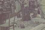 An elderly man at the ghetto market kneels by the objects he is selling with his arms extended .