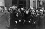 Jewish men standing among a crowd of people on a street in the Warsaw ghetto, doff their hats to the photographer, in accordance with the German order requiring Jews to remove their hats in the presence of German personnel.
