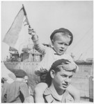 A young Jewish DP child waves a homemade flag while riding on the shoulders of an older boy at the port of Marseilles.