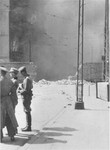 German personnel discuss matters as an apartment building razed by the SS burns during the suppression of the Warsaw ghetto uprising.