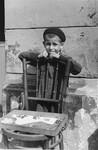 A child vendor selling candy on the street in the Warsaw ghetto, stands behind a chair waiting for customers.