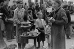 A woman drinks a cup of tea and eats a piece of bread she purchased from a vendor at an open air market in the Warsaw ghetto.