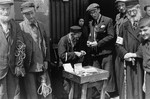 A group of people are gathered around a street vendor in the Warsaw ghetto, who is conducting business with one of them.