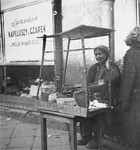 A woman seated behind a wire covered stand sells pickles on the street in the Warsaw ghetto.