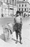 A destitute youth poses barefoot on the street in the Warsaw ghetto next to a trash can.