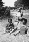 Three young children dressed in old fashioned hats pose with various household tools in the garden of the Mehoncourt children's home.