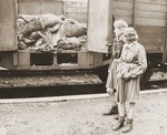 Two Jewish girls stand before a train car laden with the bodies of prisoners who died while on an evacuation transport presumably headed for Dachau concentration camp.