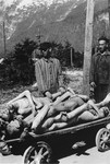 Survivors in the Ebensee concentration camp stand next to a cart stacked with the bodies of former prisoners.