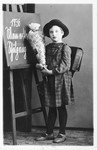 Hannelore Mansbacher on her first day of school holding a container of candy and standing near a blackboard with the caption "1936 Mein erster Schultag" (1936 my first school day).