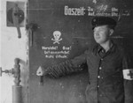 A survivor, probably Yugoslavian due to the red star on his cap, stands by the door of the Gas chamber in Dachau.