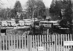 A number of Swedish Red Cross white buses, which are being used to evacuate concentration camp prisoners and transport them to Sweden, are parked in a lot behind a fence.