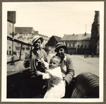 A young mother and her friend look on as her baby son pretends to drive their automobile in the main square of Nachod.