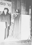 General Lucius Clay stands by a podium while Cantor Aaron Miller, draped in a prayer shawl sings at a religious convocation in an unnamed displaced persons' camp [possibly Fernwald].