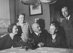 An elderly German-Jewish couple sits at their table holding hands, surrounded by their three children.