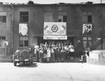 Members of Kibbutz Lochamei HaGhettaot (Ghetto Fighters) pose outside their headquarters in Berlin beneath two Hebrew banners that read: "Hashomer Hatzair" and "Kibbutz Lochamei HaGhettaot" and large portraits of Theodor Herzl and Chaim Nachman Bialik.