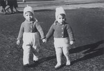 Two Jewish twins walk outside holding hands while on an outing at the lake with their parents.