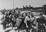 Jewish women from Subcarpathian Rus who have been selected for forced labor at Auschwitz-Birkenau, march toward their barracks carrying bed rolls after disinfection and headshaving.
