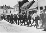 [Probably Serbian villagers from the Kozara region who have been rounded-up for deportation, marching in a column through the streets of a town.]