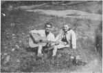 A Bosnian youth plays guitar for his girlfreind while sitting in an open field.