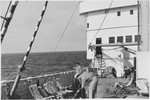 View of the bridge of the MS St. Louis.  In the foreground, a crewman cleans the deck area while a passenger standing at the railing looks out over the sea.