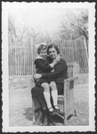 A Jewish mother and daughter pose outside their home in Moroeni, Romania, seated in a wicker chair.
