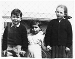 Three Czech Jewish children pose outside in front of a chain-link fence.