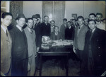 A group of men surround a "JEWCOM" official as he collects travel documents.
