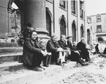 German civilian women and children sit on the steps of a largely destroyed, bomb-damaged building.