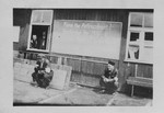 Two survivors sit outside a barrack beneath a bulletin board in the Buchenwald concentration camp.