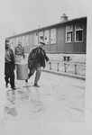 Two survivors walk past a barrack in the Buchenwald concentration camp carrying a large cannister [probably containing soup].