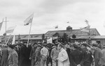 Survivors waving flags of different nationalities flock to an outdoor Jewish memorial service in the liberated Dachau concentration camp.