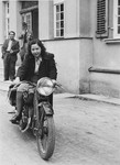 Rachel Friedman drives a motorcycle down a street in the Landsberg DP camp while her husband looks on.