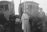 A Romani woman wearing a long white veil stands with a group, in a front of caravans decorated with flags and banners (possibly a wedding).