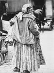 A Romani woman identified as "Liza, a Tshurara woman," stands on a city street next to a caravan.