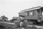 A young Romani boy sits with arms crossed, with a caravan in the background.