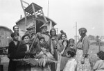 A group of Roma stand in front of their caravan.  A child with arms outstretched playfully blocks the way, and a chair is balanced aloft, above the group.