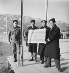 Group portrait of three Jewish displaced persons holding an Israeli flag prior to their departure for Palestine while a soldier looks on.