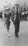 A well-dressed Jewish couple walks down a street in Vienna past a parade of Hitler Youth and hanging swastika flags.