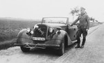 A SS officer [perhaps Max Schmidt] stands next to an automobile with an SS license plate.