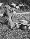A Romani man sits on the ground next to a group of cooking pots.